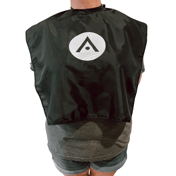 Aveda Arts Cape - Shortie, black color with screen-printed Aveda "A" logo, snap button neck, worn over a gray shirt and shorts.