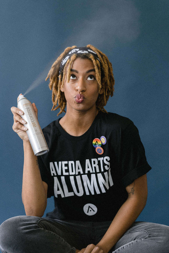 Person wearing Aveda Arts Alumni Tshirt and using hairspray, showcasing casual, relaxed style.