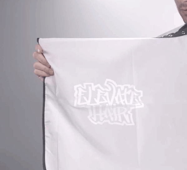 Person holding Elevate Hair white cape with logo on a grey background.