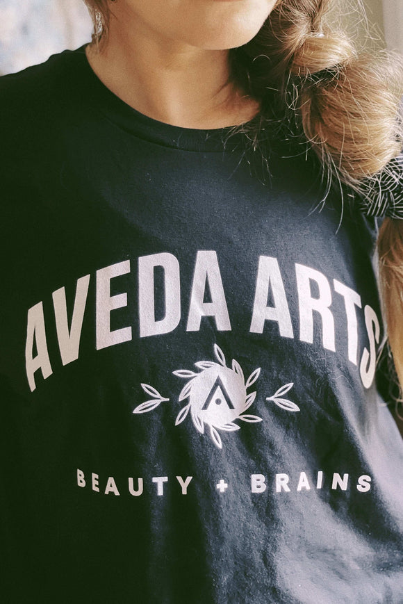 Person wearing Aveda Arts Beauty & Brains T-shirt, 100% cotton, recommending size up for a relaxed fit